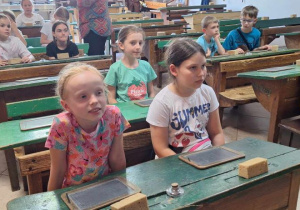 Uczniowie siedzą w ławkach starej klasy i słuchaja przewodnika