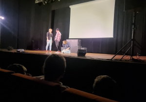 Na scenie trzy osoby, jedna osoba leży w tle ekran
