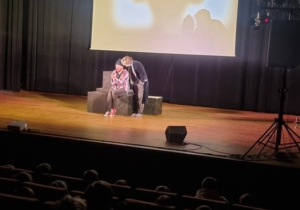 Na scenie dwie osoby, jedna osoba leży w tle ekran