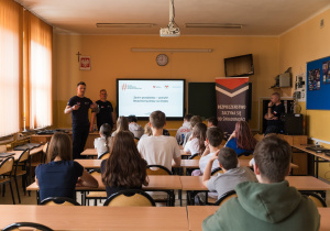Uczniowie siedzą w ławkach i słuchają prowadzących zajęcia