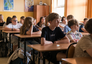 Uczniowie siedzą w ławkach i słuchają prowadzących zajęcia