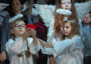 Dwa aniołki wpatrują się w czerwone serce trzymane na wyciągniętej dłoni.