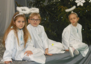 Trzy uczennice siedzą na scenie w białych strojach aniołków na czarnym tle.