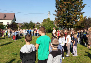 Uczniowie stoją z paniami na trawniku przed szkołą.