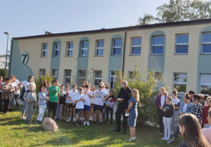 Uczniowie stoją przed szkołą i słuchają wypowiedzi z-ca dowódcy Jednostki Ratowniczo-Gaśniczej Straży Pożarnej.