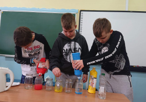 Trzech uczniów stoi przy ławce szkolnej i wlewa kolorowe płyny do słoików.