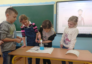 Czworo uczniów przygląda się jak koleżanka wsypuje kwasek cytrynowy do słoika z niebieskim płynem.