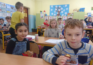 Uczniowie siedzą w ławkach i jedzą pączki.