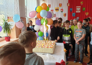 Uczniowie stoją na korytarzu, na stoliku jest tort, obok kolorowe balony.