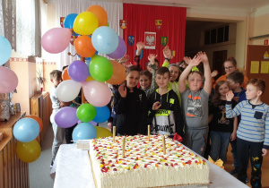 Uczniowie stoją na korytarzu szkolnym obok stolika na którym jest tort.