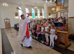 Ksiądz święci przybory szkolne, uczniowie stoją w ławkach.