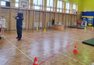 Dwoje uczniów przygotowuje się do startu, obok stoi policjant, w tle tor przeszkód.