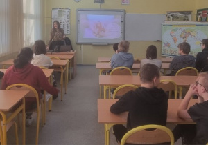 Uczniowie słuchają wykładu na temat profilaktyki w oddali stoi pani prowadząca zajęcia.