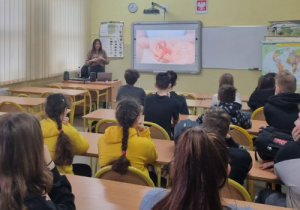 Uczniowie słuchają wykładu na temat profilaktyki w oddali stoi pani prowadząca zajęcia.