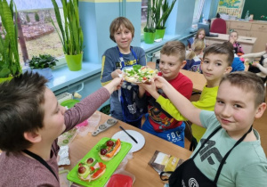 Dzieci siedzą przy ławkach, na stołach leżą zdrowe kanapki.