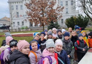 Uczniowie stoją w grupie na placu przed UM i słuchają wyjaśnień nauczyciela