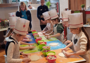 Uczniowie stoją przy stole i robią pizzę.