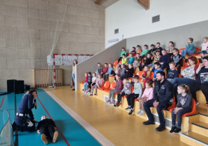 Uczniowie siedzą na trybunach i słuchają policjantki, na podłodze leży uczeń.