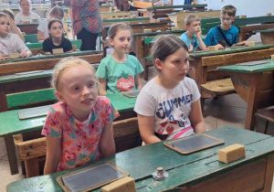 Uczniowie siedzą w ławkach starej klasy i słuchaja przewodnika