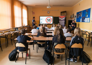 Uczniowie siedzą w ławkach i słuchają prowadzących zajęcia