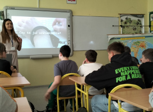 Uczniowie siedzą w ławkach i słuchają prowadzącej.