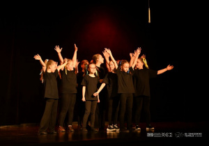 Duża grupa uczniów stoi na scenie z wyciągniętymi ramionami