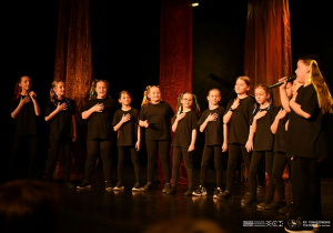 Dwanaście uczennic stoi na scenie i śpiewa