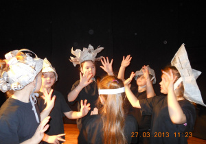 Grupa dziewczynek ubranych w czarne stroje i kapelusze z gazet tańczy w kole wymachując rękoma, twarze zwrócone do środka koła.